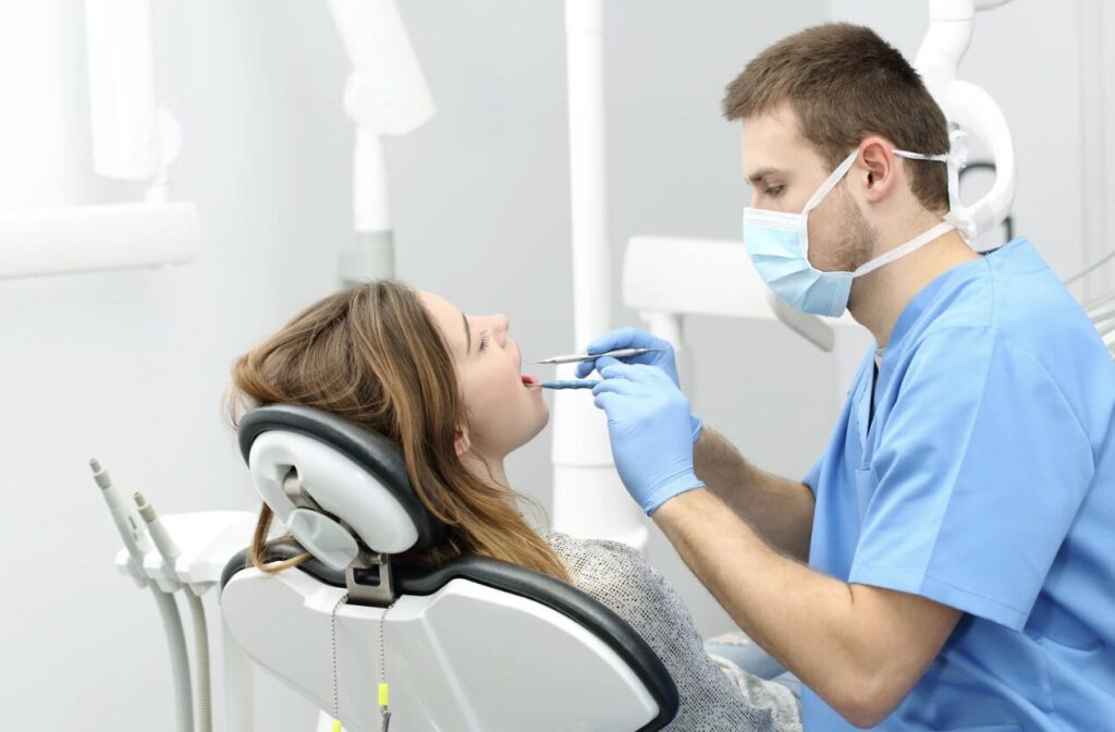 Dentist in blue scrubs and mask uses tools to examine a woman's open mouth in a dental chair.