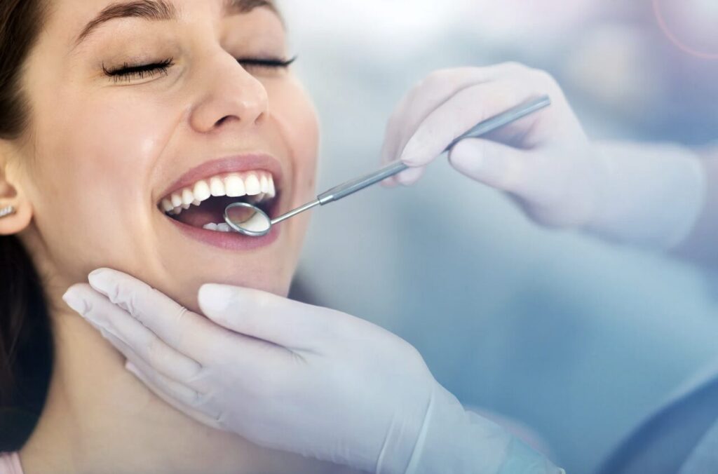 Woman smiling during a dental exam as a dentist uses a mirror and tool to inspect her teeth.