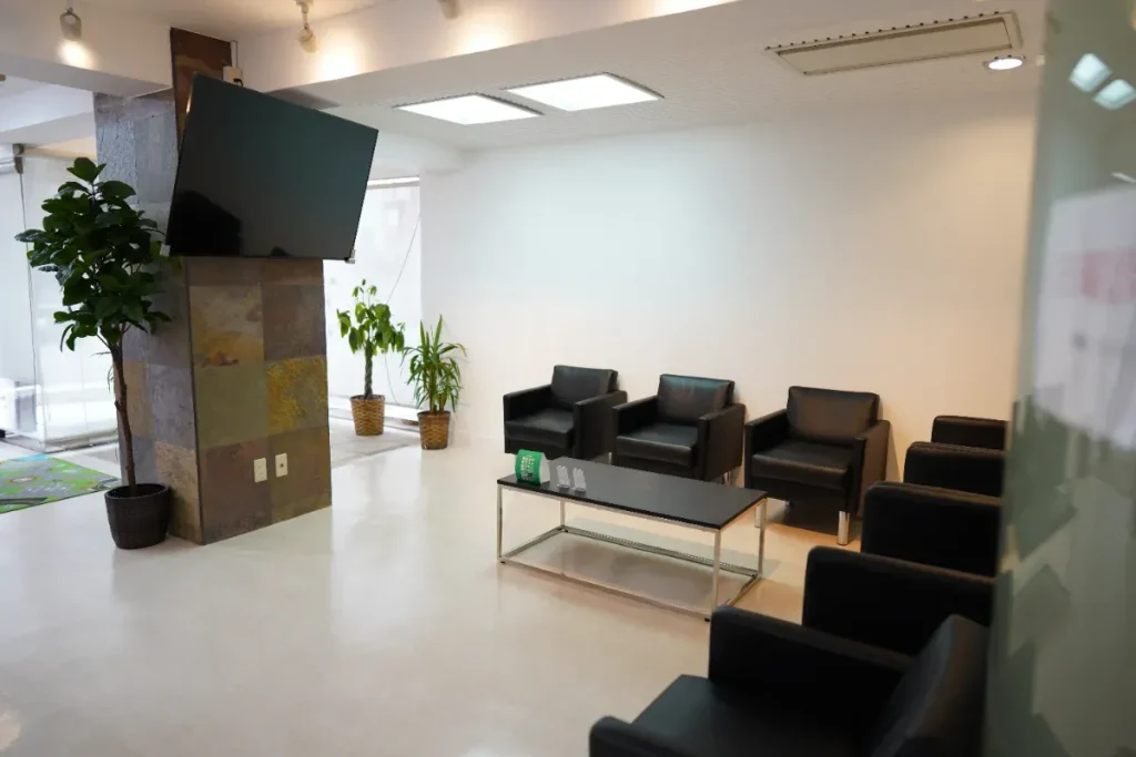 Modern waiting area with black leather chairs arranged around a dark coffee table, white walls, and a large flat-screen TV mounted on a stone-clad pillar; several potted plants nearby.