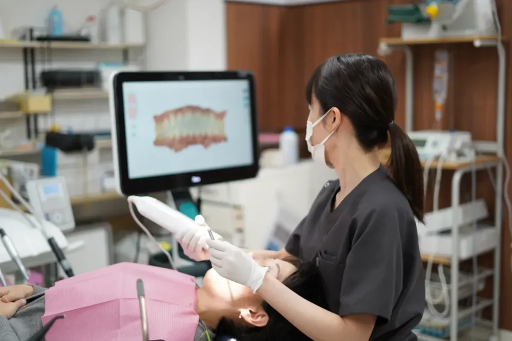 Dental clinician wearing a mask and gloves treats a patient with dental instruments, monitor showing a dental image in the background.