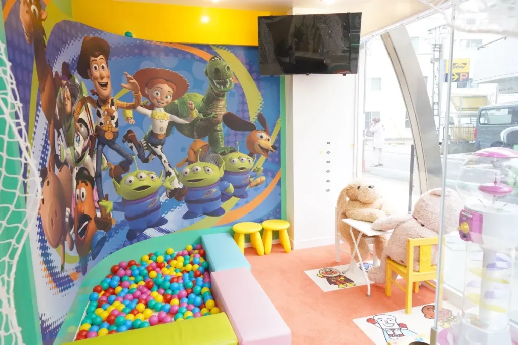 Bright kids' play area with a multicolored ball pit, pastel foam seating, and a large cartoon mural on the wall behind it.