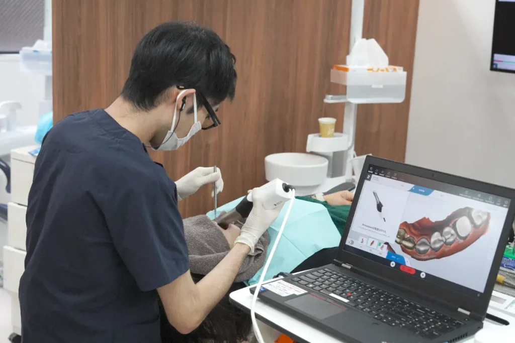 Dentist in scrubs and mask treats a patient while a laptop screen shows a 3D dental model.
