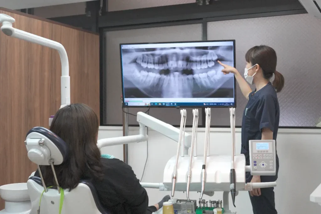 Patient seated in a dental chair while a clinician points to a panoramic tooth X-ray displayed on a monitor. There are dental tools nearby for treatment.